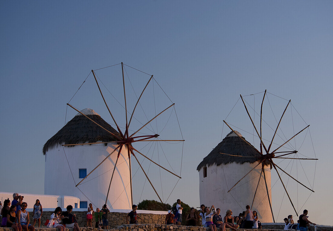 Menschen beobachten den Sonnenuntergang unter den alten Windmühlen; Mykonos-Stadt, Mykonos, Kykladen, Griechische Inseln, Griechenland