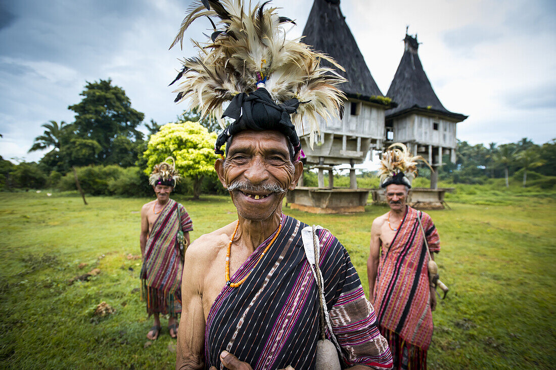 Gruppe von Männern in traditioneller Kleidung mit heiligen Häusern im Hintergrund; Distrikt Lospalmos, Timor-Leste.