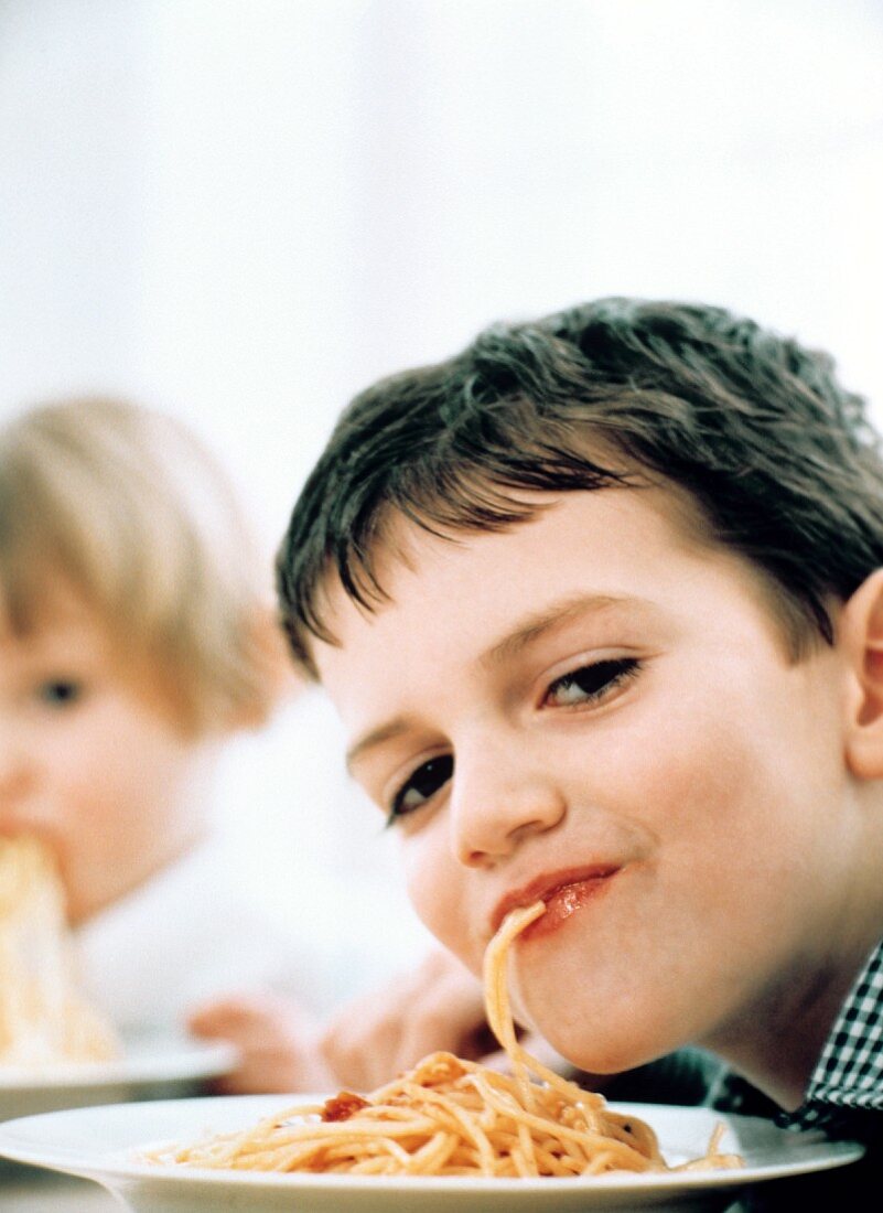 Kleiner Junge isst Spaghetti, im Hintergrund ein Mädchen