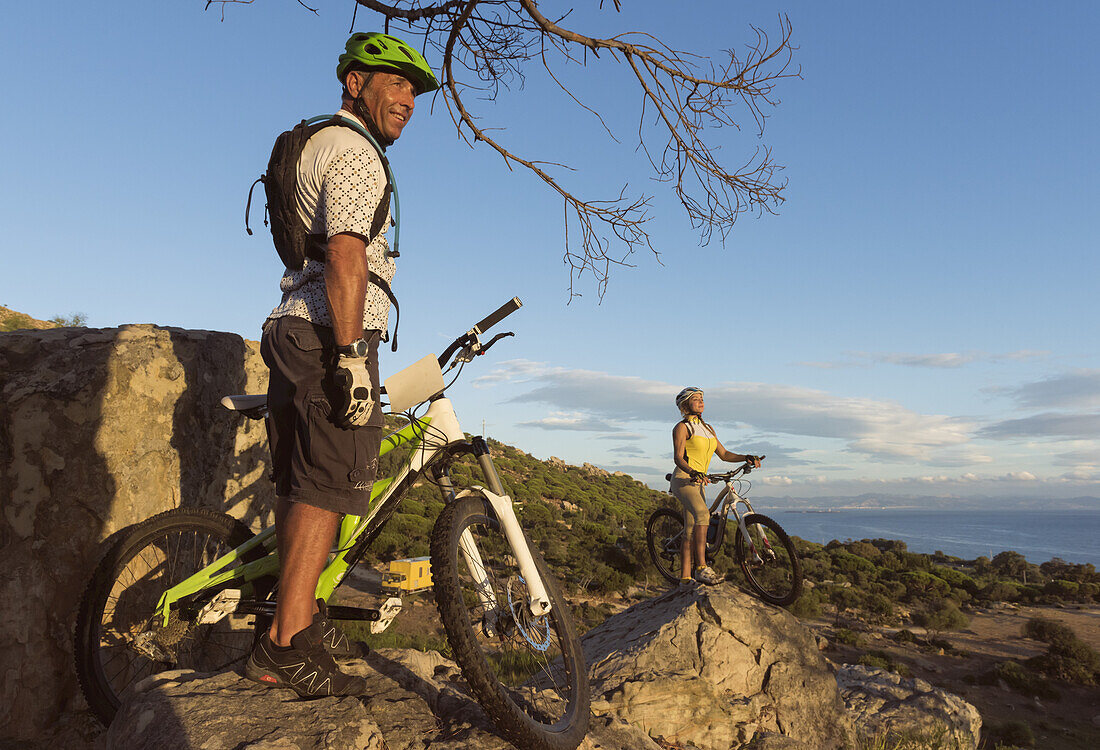 A Man And Woman Standing With Their Bicycles On A Rocky Ledge Overlooking A View Of The Landscape; Tarifa, Cadiz, Andalusia, Spain