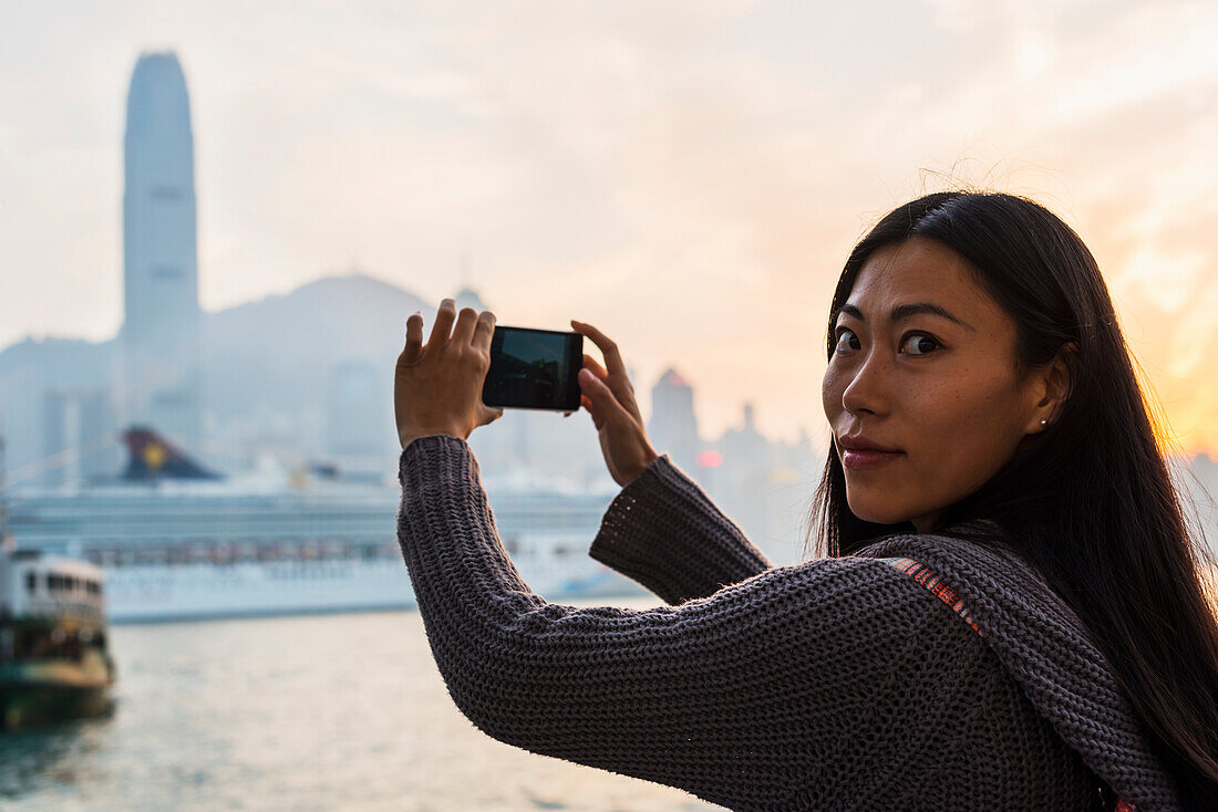 Eine junge Frau fotografiert Gebäude auf der anderen Seite des Wassers, Kowloon; Hongkong, China