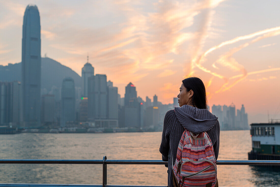 A Young Woman At The Waterfront With A View Of The Skyline At Sunset, Kowloon; Hong Kong, China