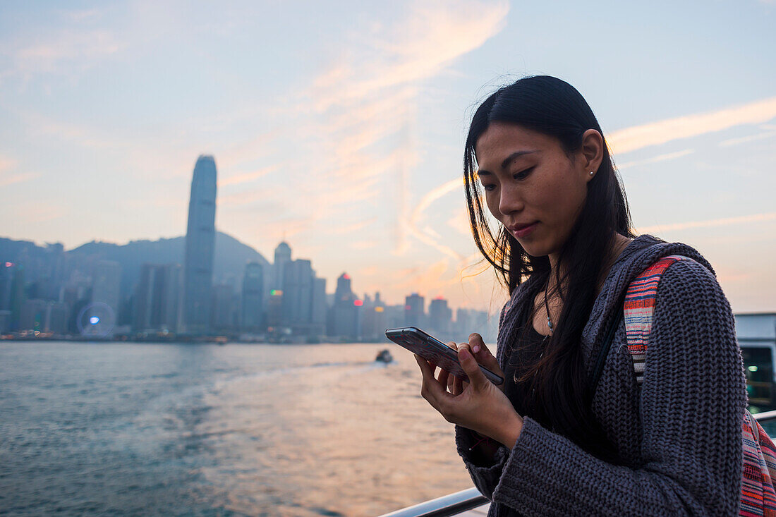 Eine junge Frau prüft ihr Handy an der Waterfront mit Blick auf die Skyline bei Sonnenuntergang, Kowloon; Hongkong, China.