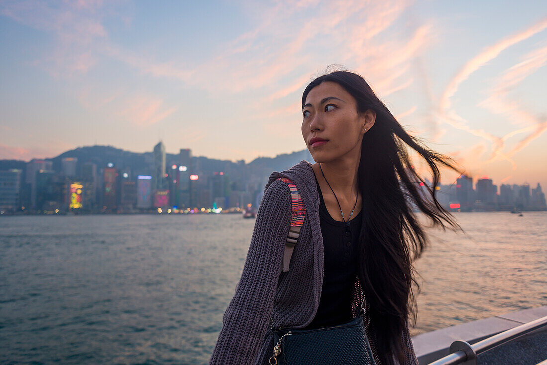 Eine junge Frau am Ufer, die bei Sonnenuntergang auf dem Geländer sitzt, mit der Skyline im Hintergrund, Kowloon; Hongkong, China.