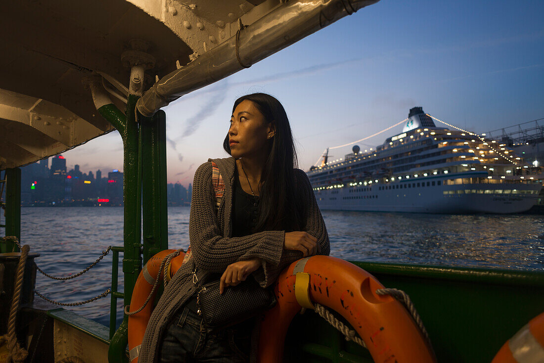 Junge Frau am Wasser bei Sonnenuntergang mit einem Kreuzfahrtschiff im Hintergrund, Kowloon; Hongkong, China.