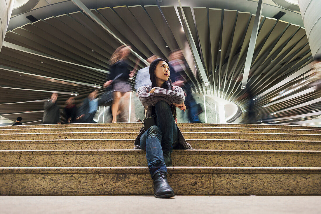 A Young Woman Sitting On Steps With A Modern Building And Pedestrians In The Background, Kowloon; Hong Kong, China