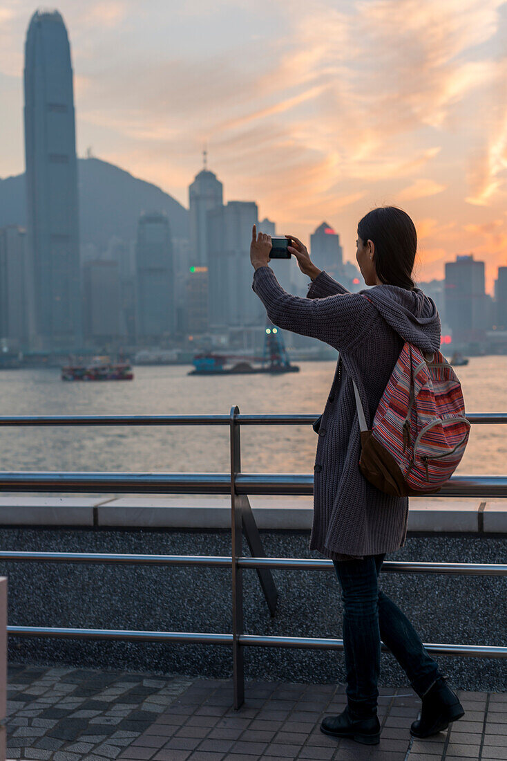 Eine junge Frau fotografiert mit ihrer Kamera den Hafen und die Skyline von Hongkong bei Sonnenuntergang, Kowloon; Hongkong, China
