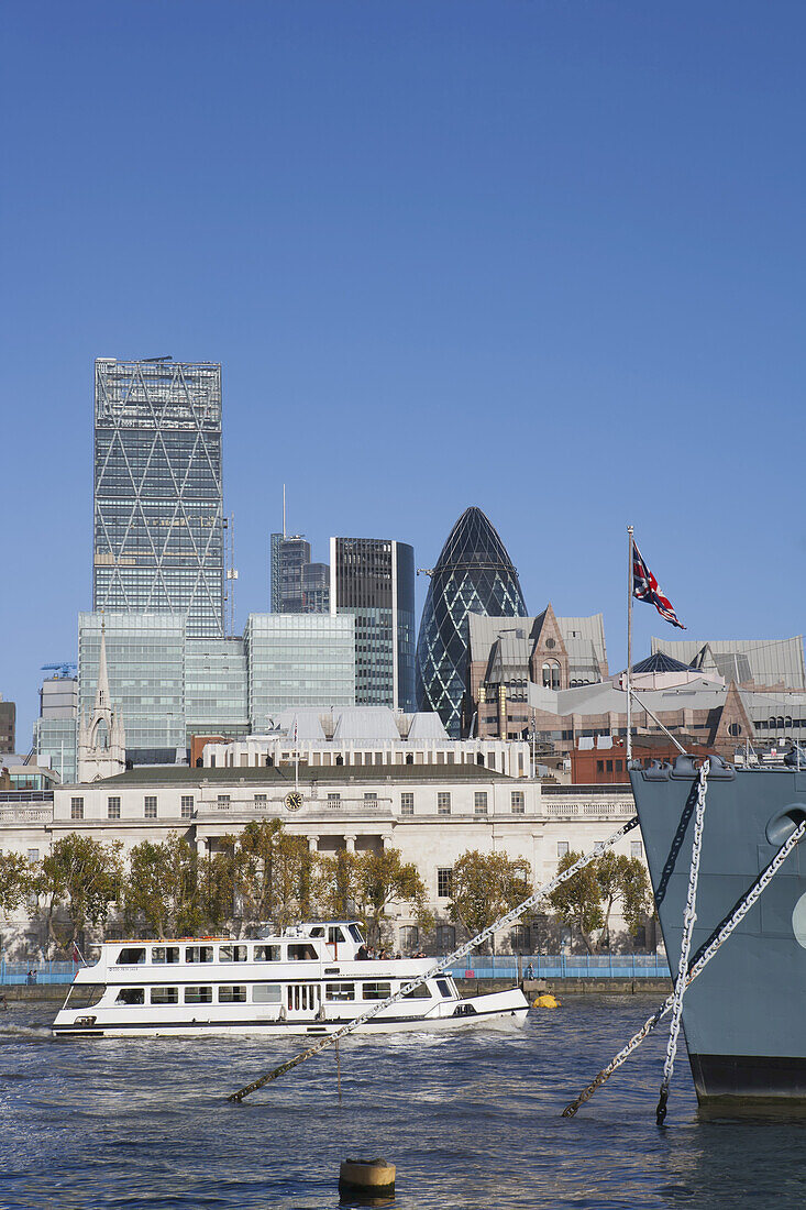 Ein Touristenboot fährt am Bug des Kriegsschiffs HMS Belfast vorbei, das an der Themse liegt und Teil des Imperial War Museum ist, mit Wolkenkratzern in der Londoner City, darunter die Gurke von Norman Foster; London, England.