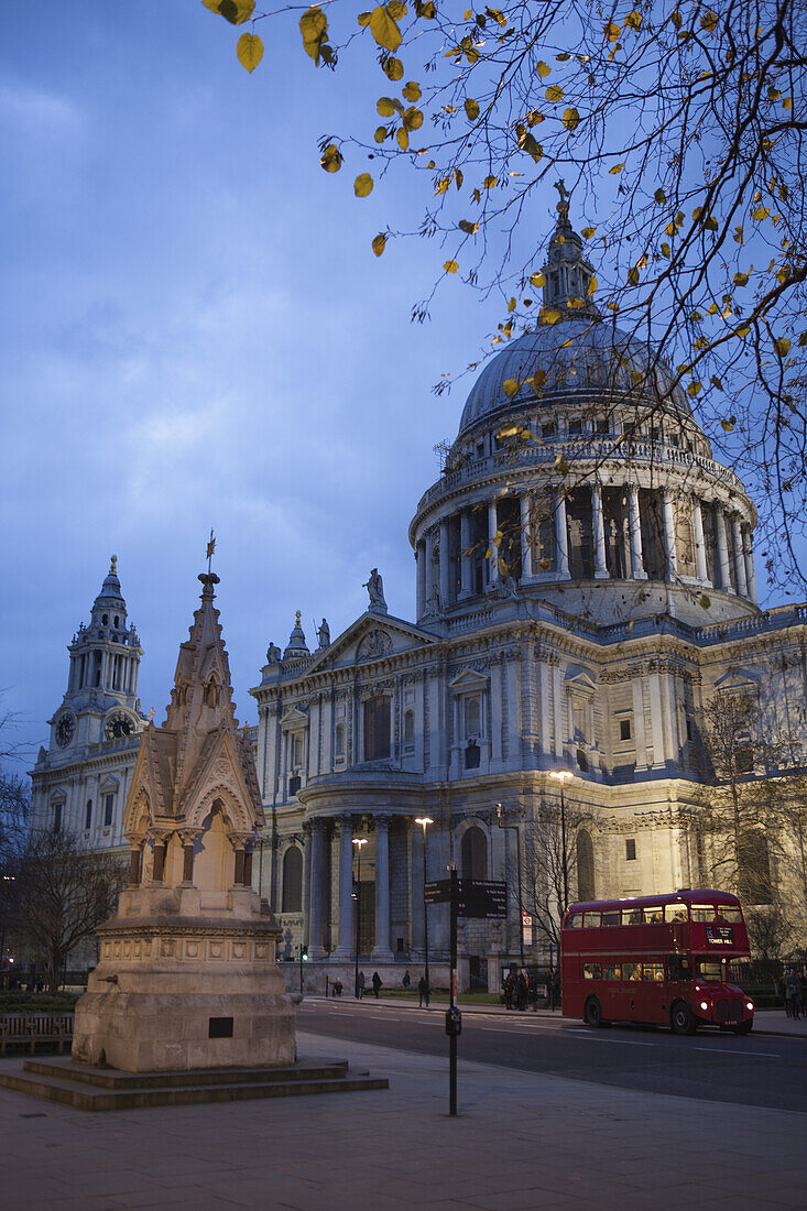 A Red London Route Master Double-Decker Bus Passes St Paul's Cathedral At Dusk, With The St Lawrence Jewry Memorial Fountain In The Foreground; London, England