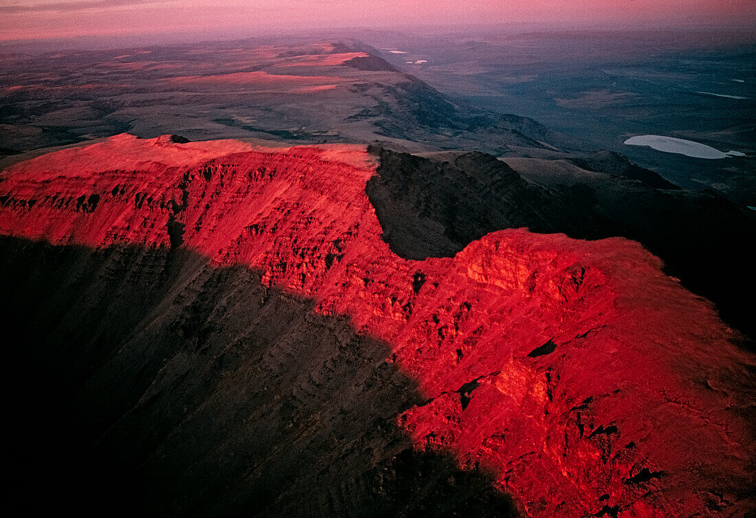 Blazing sunset leaves in shadow the famous gap in Kiger Gorge, atop Oregon's Steens Mountain. Steen's Mountain Wilderness is 'the largest fault-block mountain in the northern Great Basin'. The aerial view shows a forty mile long escarpment in southeastern Oregon has a notch cut out of the top and drops abruptly to the dry Alvord Desert, 5,500 feet below. Bulldozing down to basalt, Ice Age glaciers carved our huge gorges out of the Great Basin's largest fault block mountain. Beyond, Steens's east face plummets a vertical mile; Oregon, United States of America