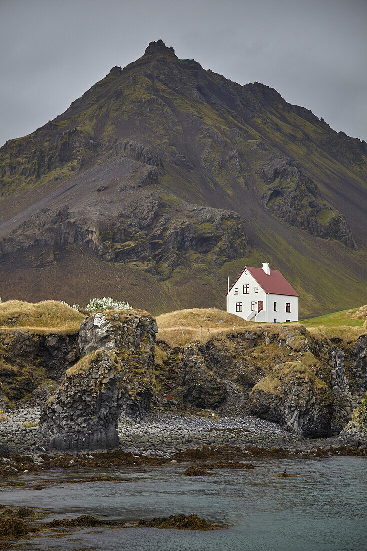 Lone white house and rugged mountain with lava cliffs along the seashore at Arnastapi, Snaefellsnes, western Iceland; Arnastapi, Iceland