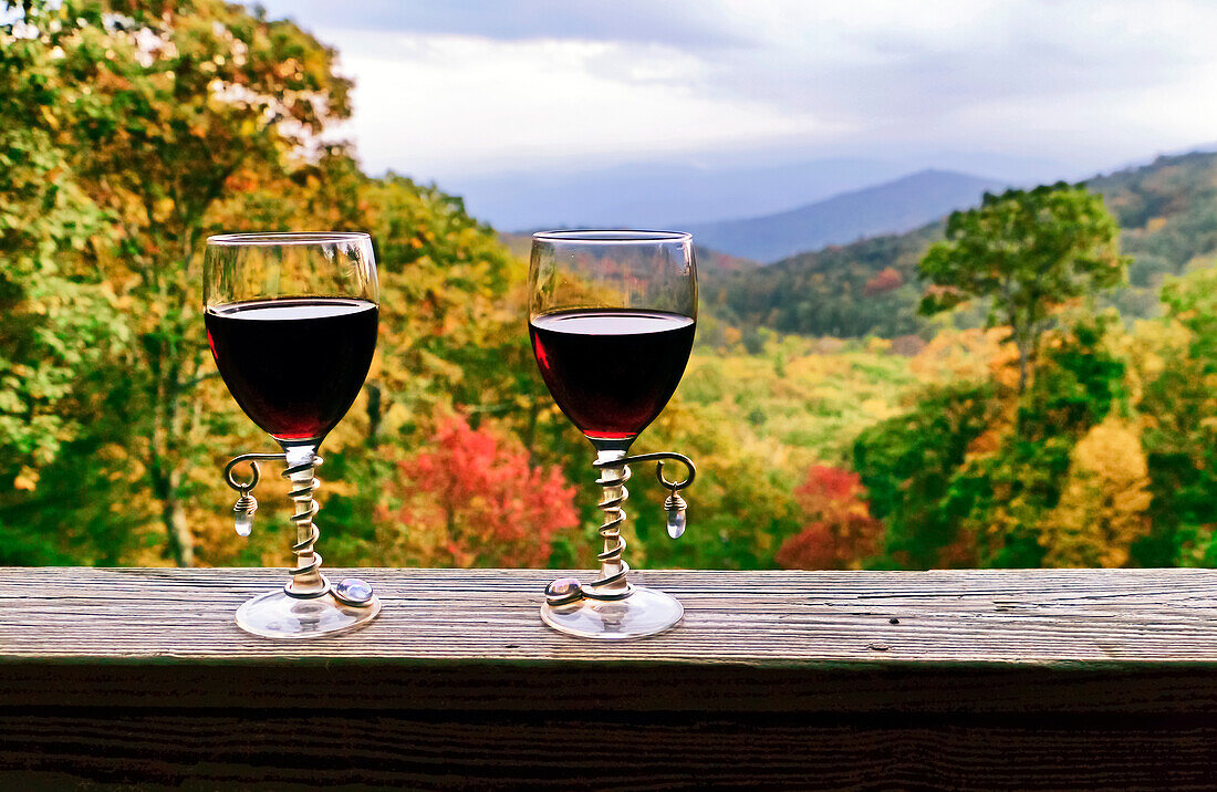 Two decorative glasses of red wine sit on a wooden railing above a mountain view in autumn; Fairview, North Carolina, United States of America