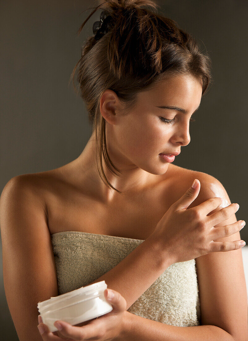 Young woman wrapped in a towel holding cream jar and rubbing her shoulder with body lotion