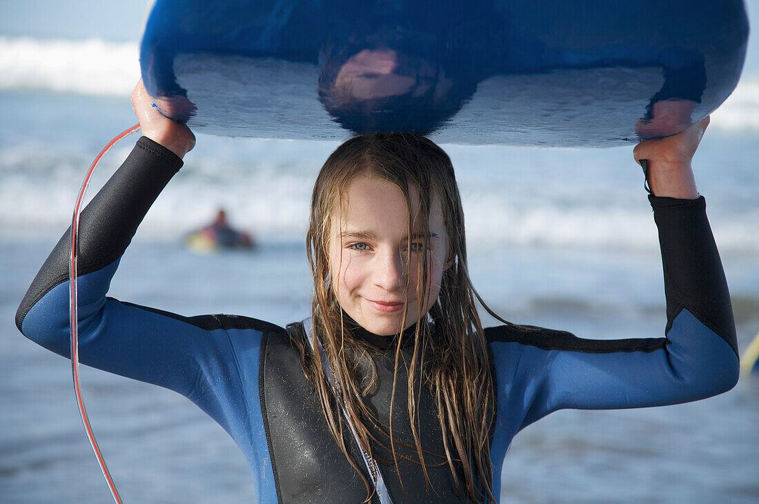 Lächelndes und nasses Mädchen, das ein Surfbrett auf dem Kopf hält und aus dem Meer kommt