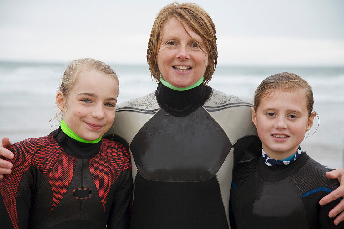 Woman and two girls wearing surfing wetsuits standing on a beach smiling
