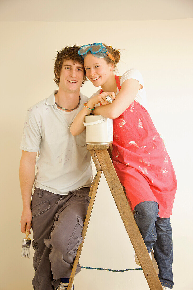 Lächelndes junges Paar lehnt mit dem Gesicht zueinander auf einer Leiter und hält Pinsel