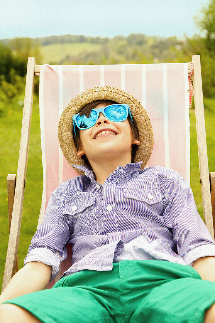 Lächelnder Junge mit Strohhut und Sonnenbrille auf einem Liegestuhl sitzend