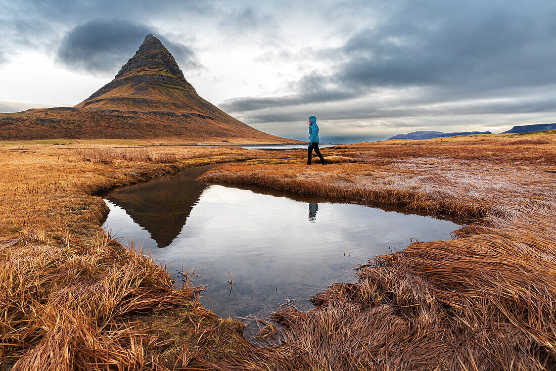 Frau geht vor dem berühmten Berg Kirkjufell spazieren, der sich im Wasser eines kleinen Sees spiegelt, Snaefellsnes Halbinsel, Westisland, Island, Polarregionen