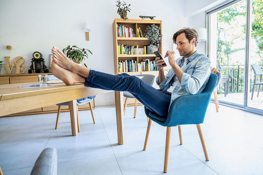 Young adult man resting feet while working with smart phone at home
