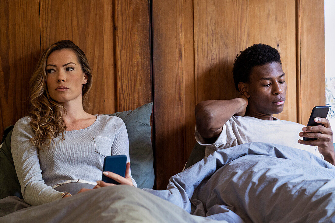 Adult couple lying on bed with upset look on faces while using smart phone