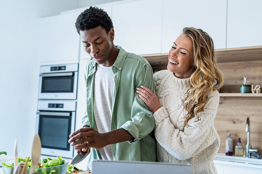 Adult woman embracing boyfriend while cutting vegetables at kitchen counter