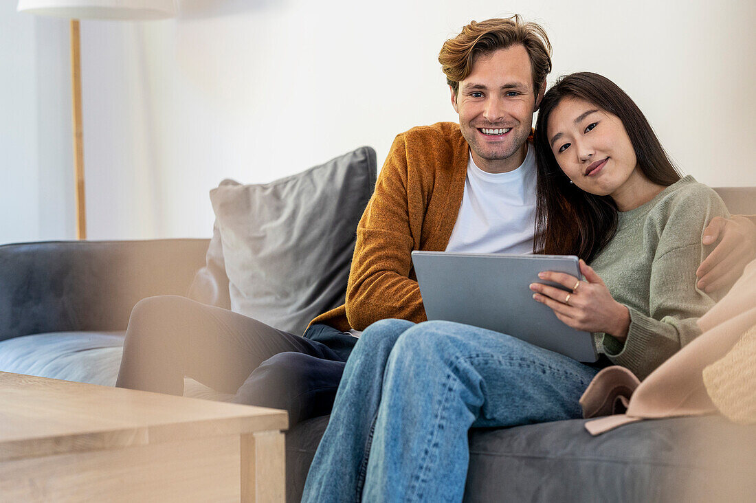 Erwachsenes Paar schaut in die Kamera, während es auf dem Sofa sitzt und ein digitales Tablet benutzt