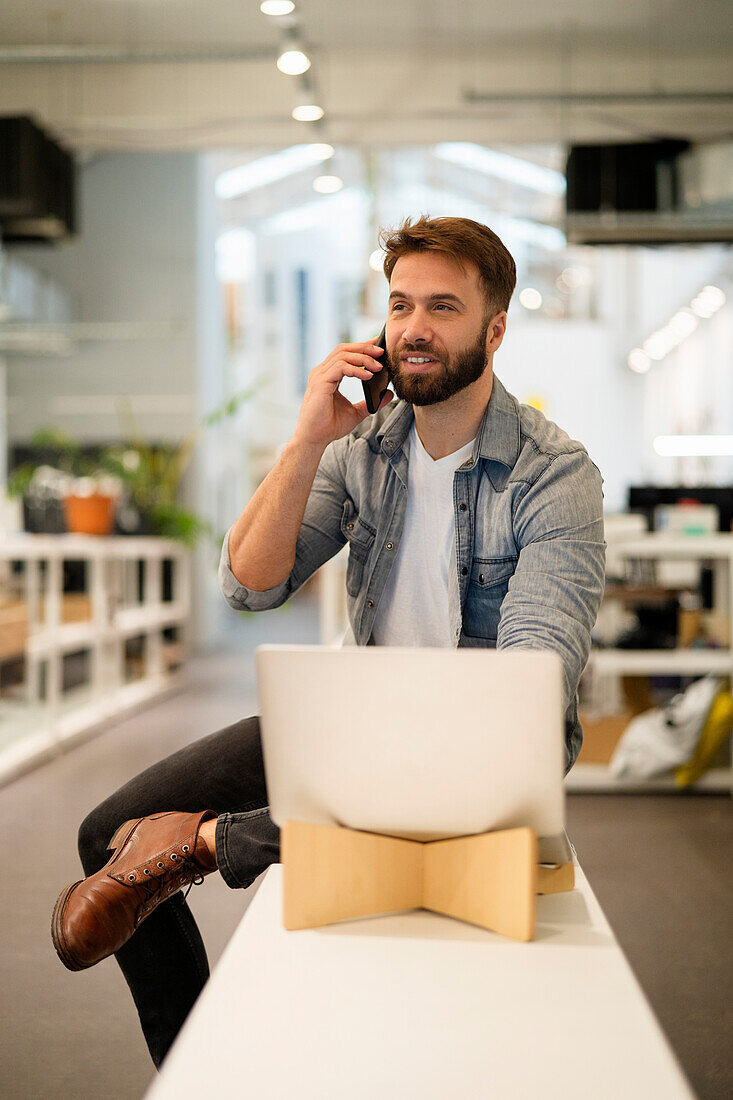Male entrepreneur sitting on table while talking on smart phone
