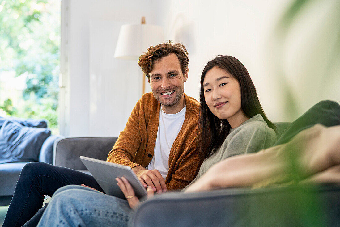 Adult couple looking at the camera while sitting on sofa using digital tablet