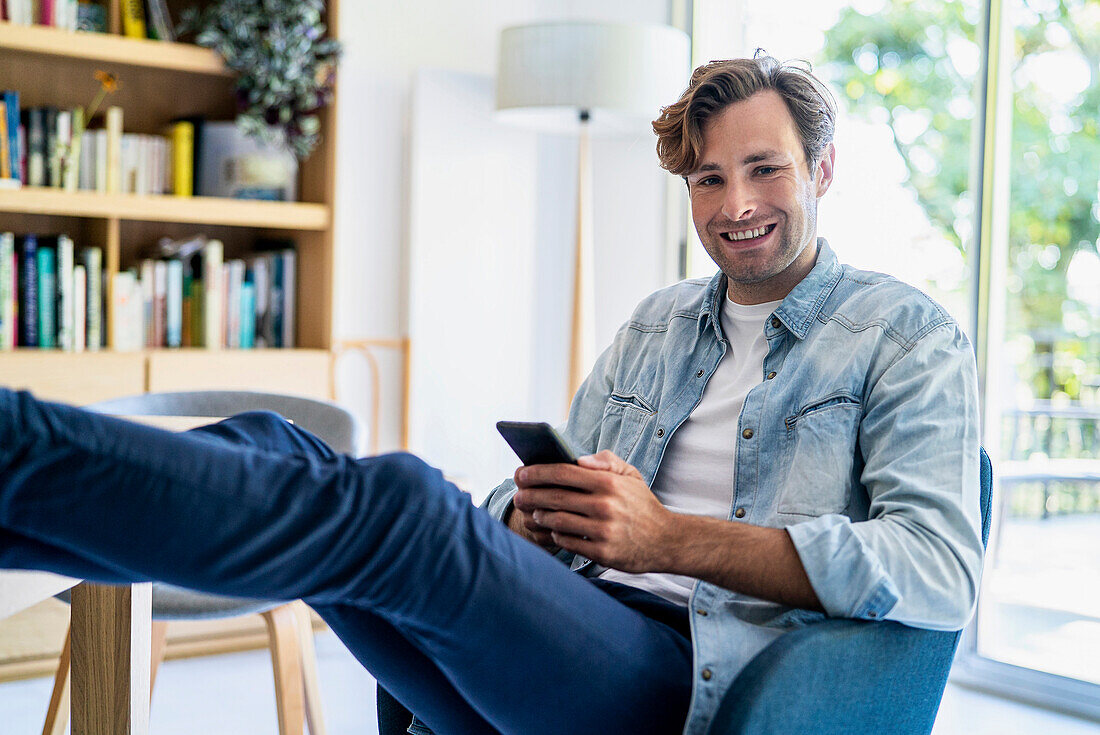 Geschäftsmann benutzt ein Smartphone, während er auf einem Stuhl sitzt und die Füße auf den Tisch legt