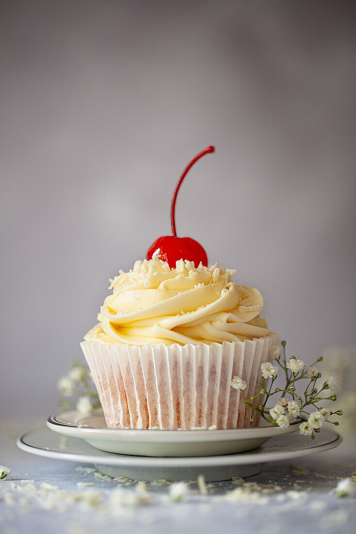 A white chocolate cupcake topped with buttercream, grated white chocolate and a stemmed maraschino cherry. Presented on a plate with white flowers.