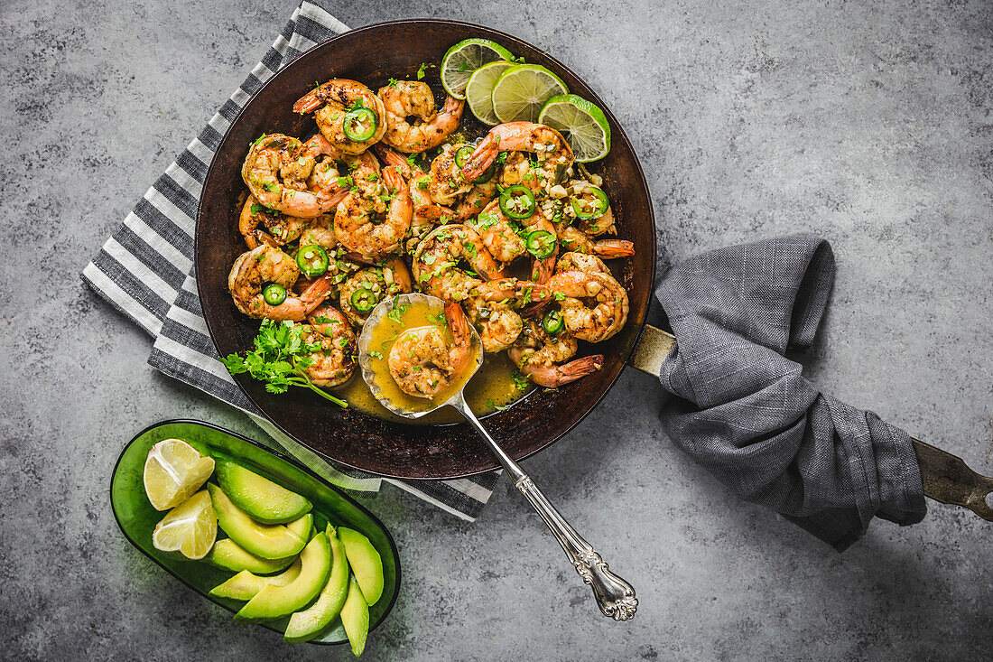 Prawns with chilli, lime and coriander in butter sauce, carbon steel pan, with avocado slices on grey linen napkins and silver spoon
