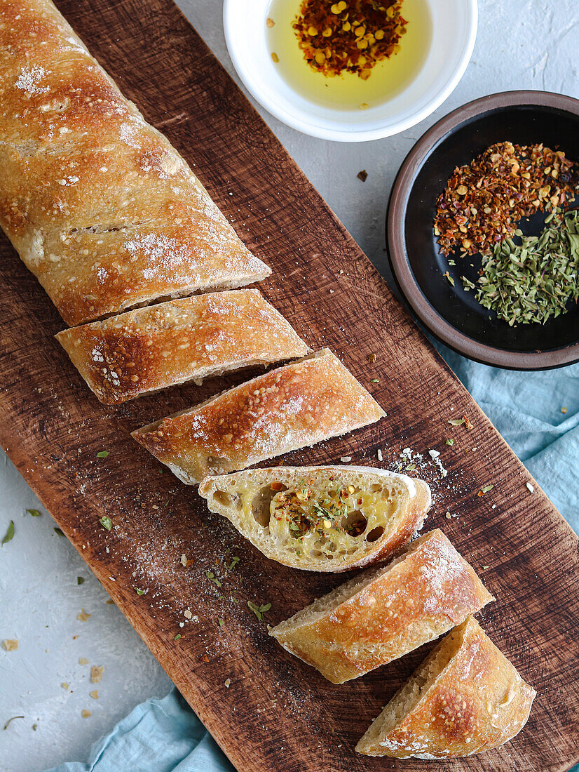 Aufgeschnittenes Baguettebrot mit Olivenöl, Oregano und Chiliflocken auf einem Holzschneidebrett