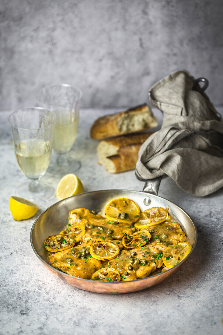 Chicken, lemon slices and capers in a copper pan with a napkin on the handle, bread and white wine