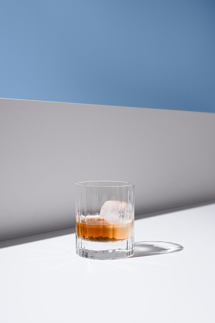 Transparent glass of cold refreshing bourbon with ice cube placed on white surface near white wall