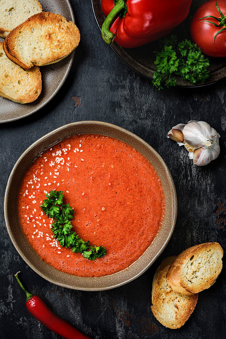 Gazpacho garnished with parsley in a wavy-edged plate. On the table are red peppers, tomatoes, garlic and croutons. View from above