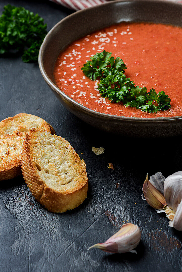 Gazpacho garniert mit Petersilie in einem Teller mit gewelltem Rand. Auf dem Tisch liegen Knoblauch und Croutons