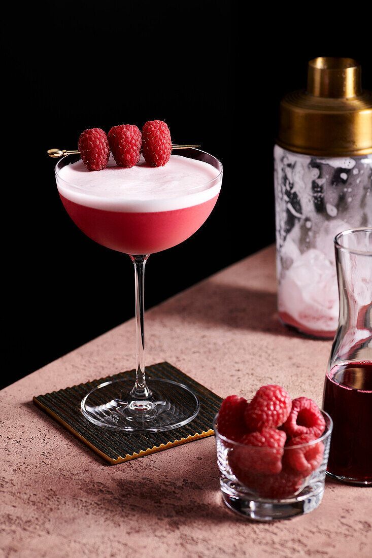Clover club cocktail in a tall coupe glass garnished with three raspberries on a gold pick.