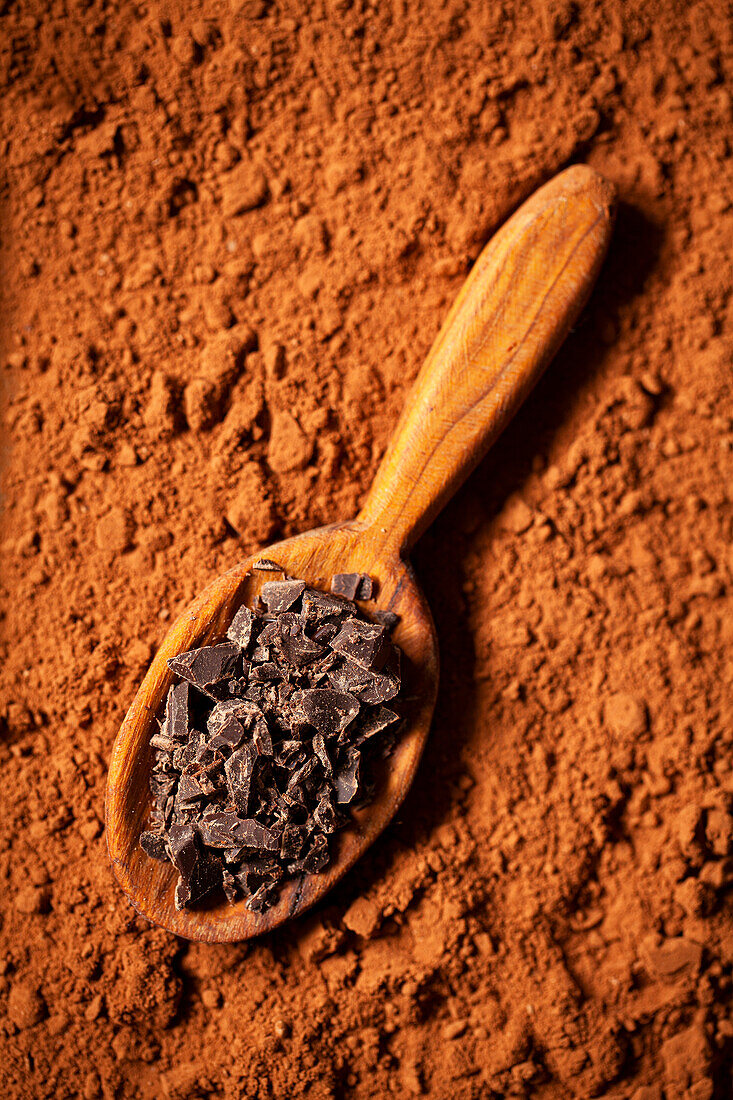 Dark chocolate chips on a wooden spoon resting on a layer of cocoa powder