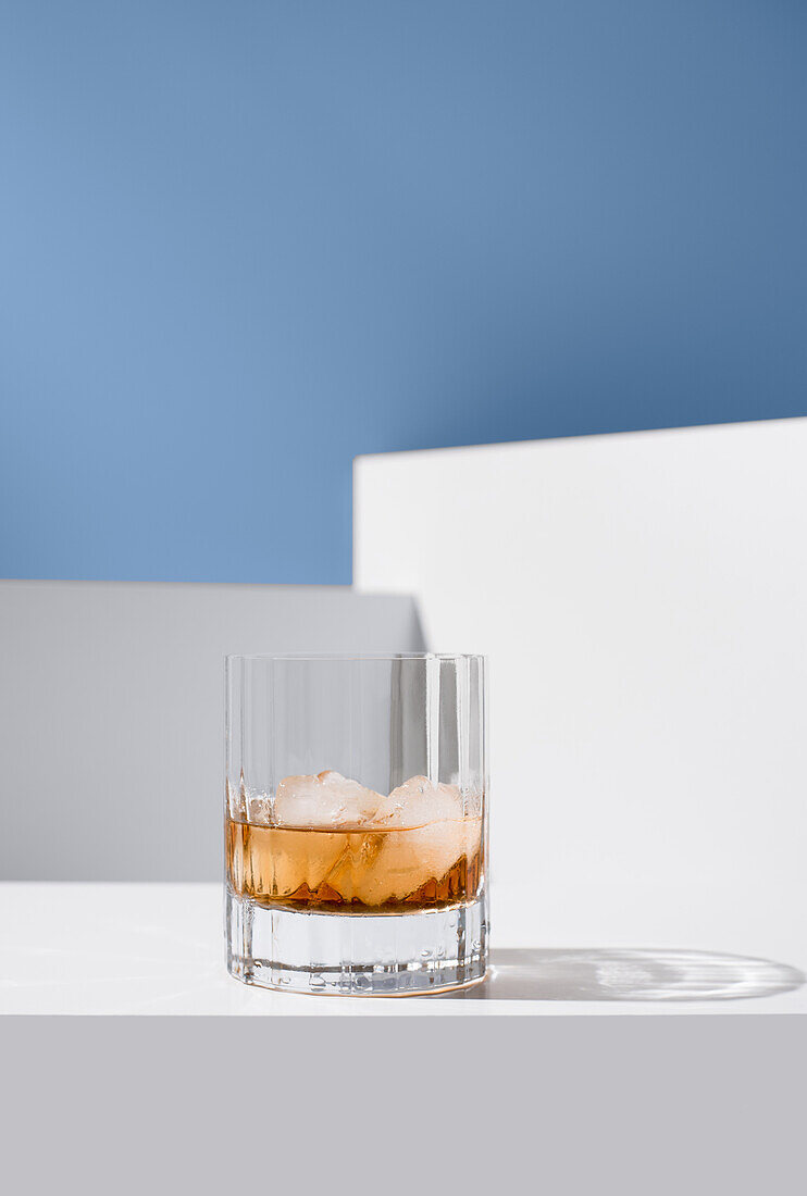 Close up of transparent glass of whiskey with ice cubes placed on white surface against white walls