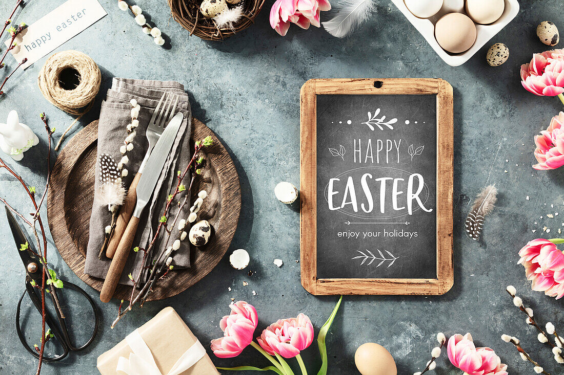 Ostertisch mit Frohe Ostern Text auf Kreidetafel, Frühlingsblumen und Besteck auf dunkelblauem Hintergrund Draufsicht Flat Lay