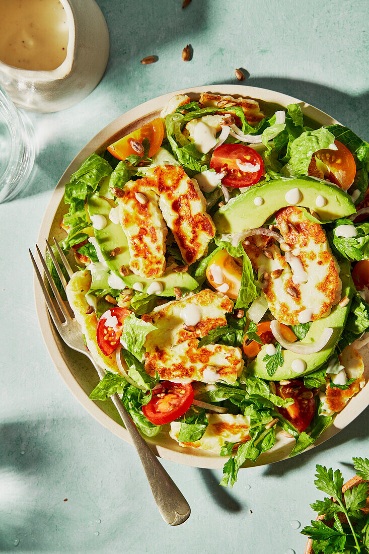 Halloumi avocado tomato salad with sunflower seeds, yoghurt dressing, herbs and water on a green background with shade