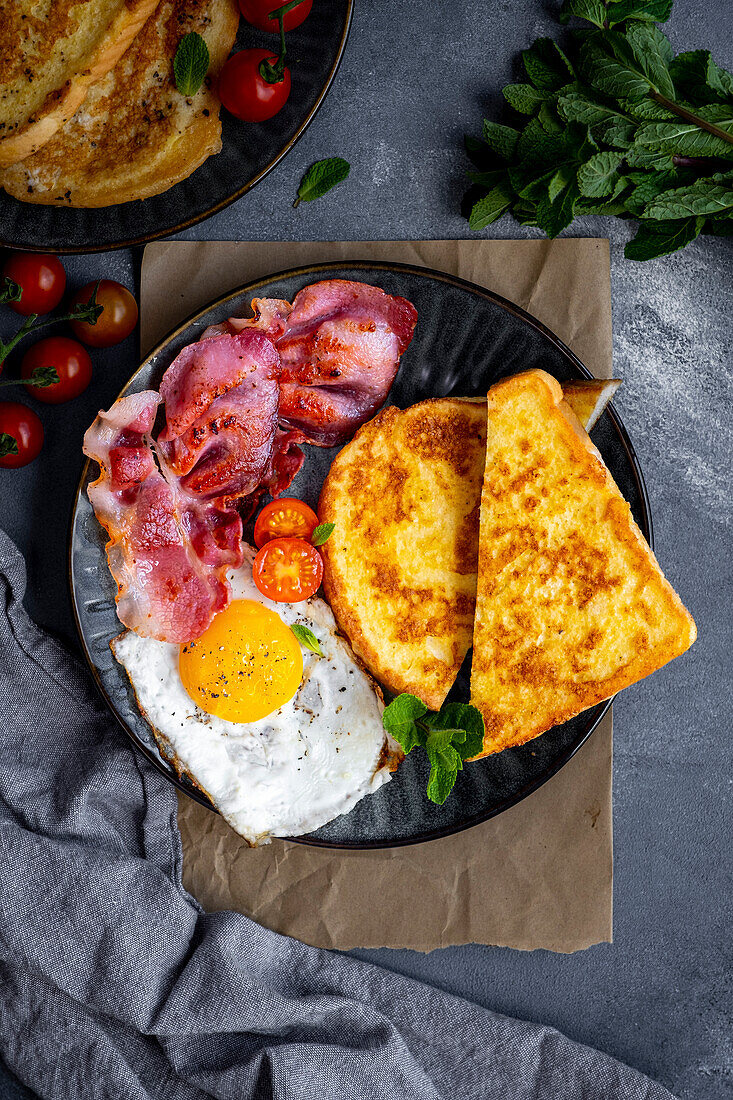 A breakfast plate with French toast, fried egg and bacon, garnished with fresh mint leaves and cherry tomatoes