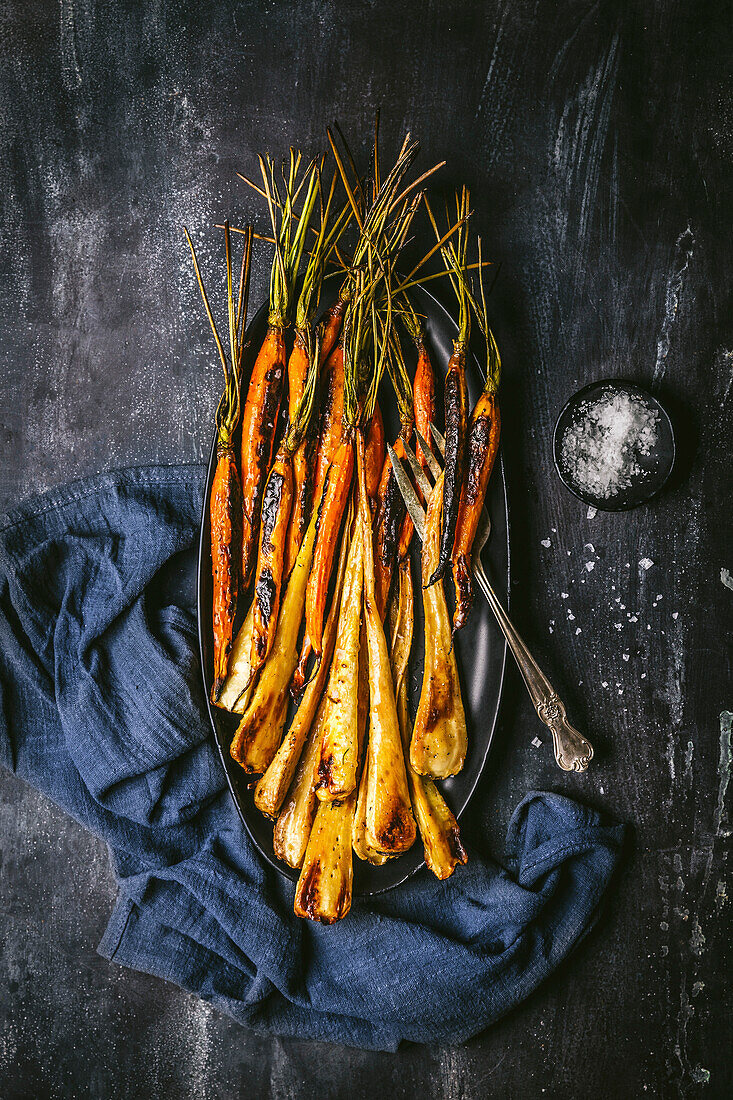 Roasted, glazed carrots and parsnips on a dark oval platter with serving fork and blue linen napkin on a dark background
