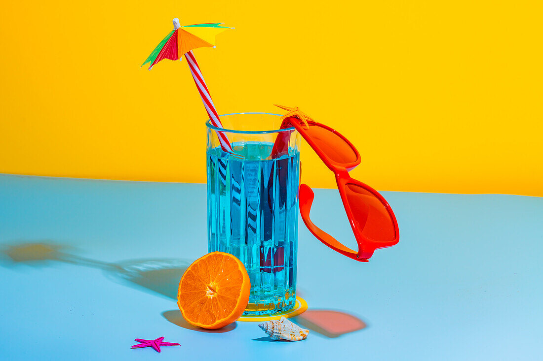 Front view of a composition of a glass with blue liquid and a straw with stylish red glasses against a light blue and yellow background in the studio