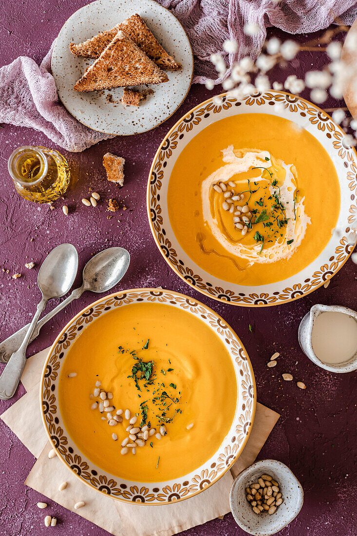 Two bowls of pumpkin soup, served with brown toast and garnished with cream, pine nuts and herbs on a purple background