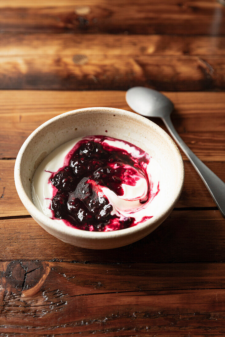 Bowl of yoghurt and blackberry compot on wooden table with silver spoon.