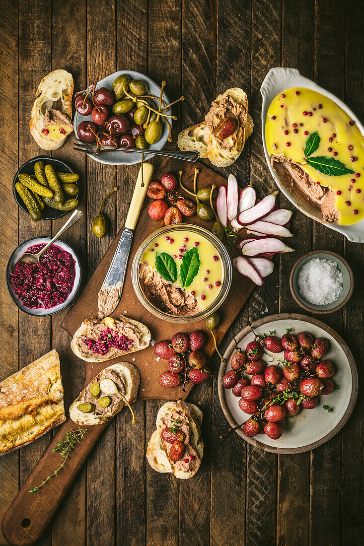 Pâté spread in a jar on a wooden board with roasted grapes, radishes, gherkins and baguette