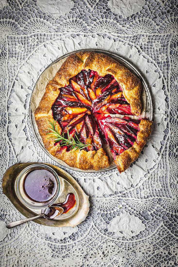 Rustic plum galette, sliced, with red wine-caramel sauce and a sprig of fresh rosemary on a background of intertwined lace