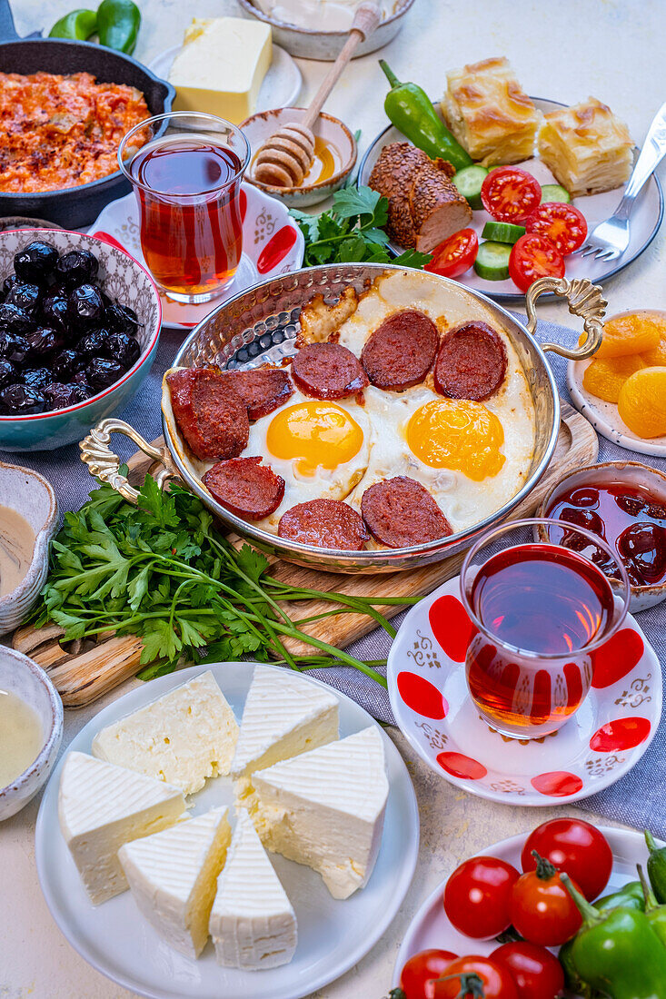 Türkischer Frühstückstisch aus der Vorderansicht. Eier und Sujuk in einer Kupferpfanne. Oliven, Käse und türkischer Tee in Tulpengläsern drumherum