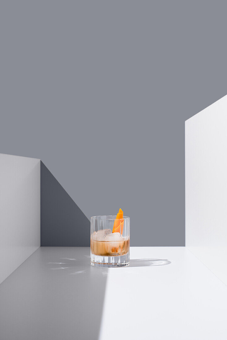Kristallklares Glas gefüllt mit frischem altmodischem Cocktail, garniert mit Orangen und Eiswürfeln auf weißer Fläche zwischen weißen Wänden vor grauem Hintergrund